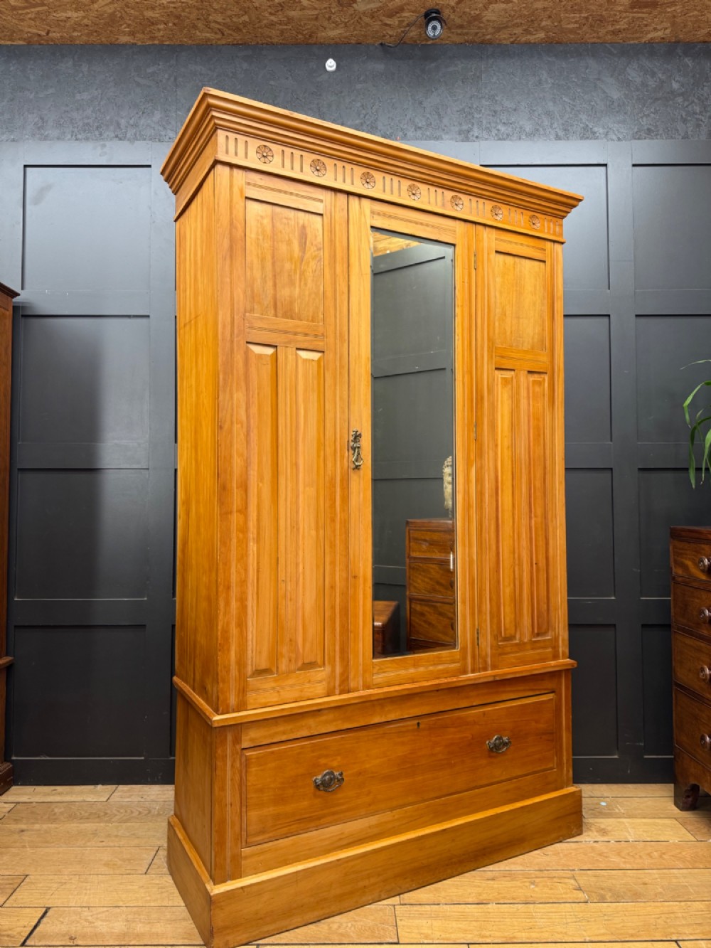 A tall Edwardian satinwood wardrobe featuring a central mirrored door, panelled side doors, a carved rosette frieze, and a wide drawer at the base.