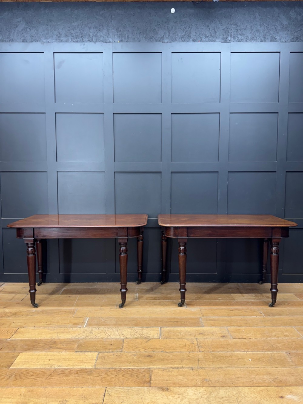 A matching pair of dark mahogany side tables featuring turned legs and brass castors standing against a panelled wall.