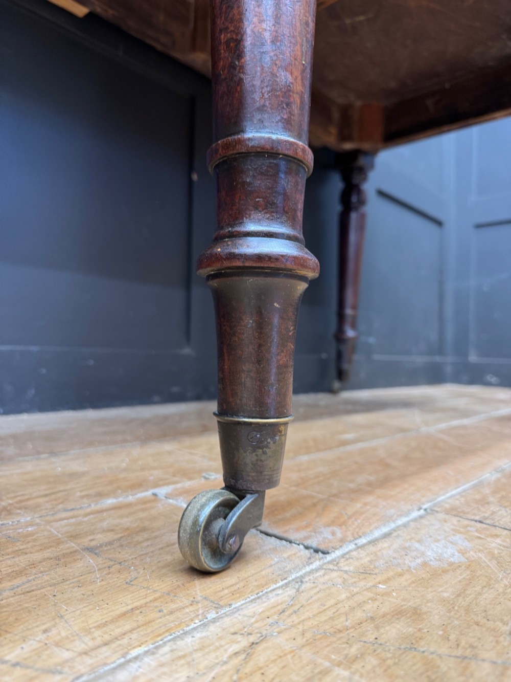A matching pair of dark mahogany side tables featuring turned legs and brass castors standing against a panelled wall. View VII