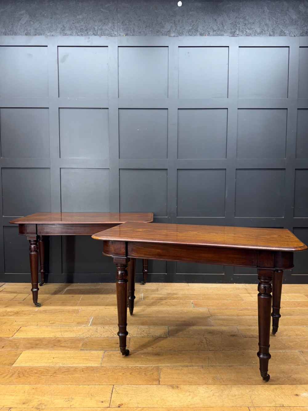 A matching pair of dark mahogany side tables featuring turned legs and brass castors standing against a panelled wall. View VIII
