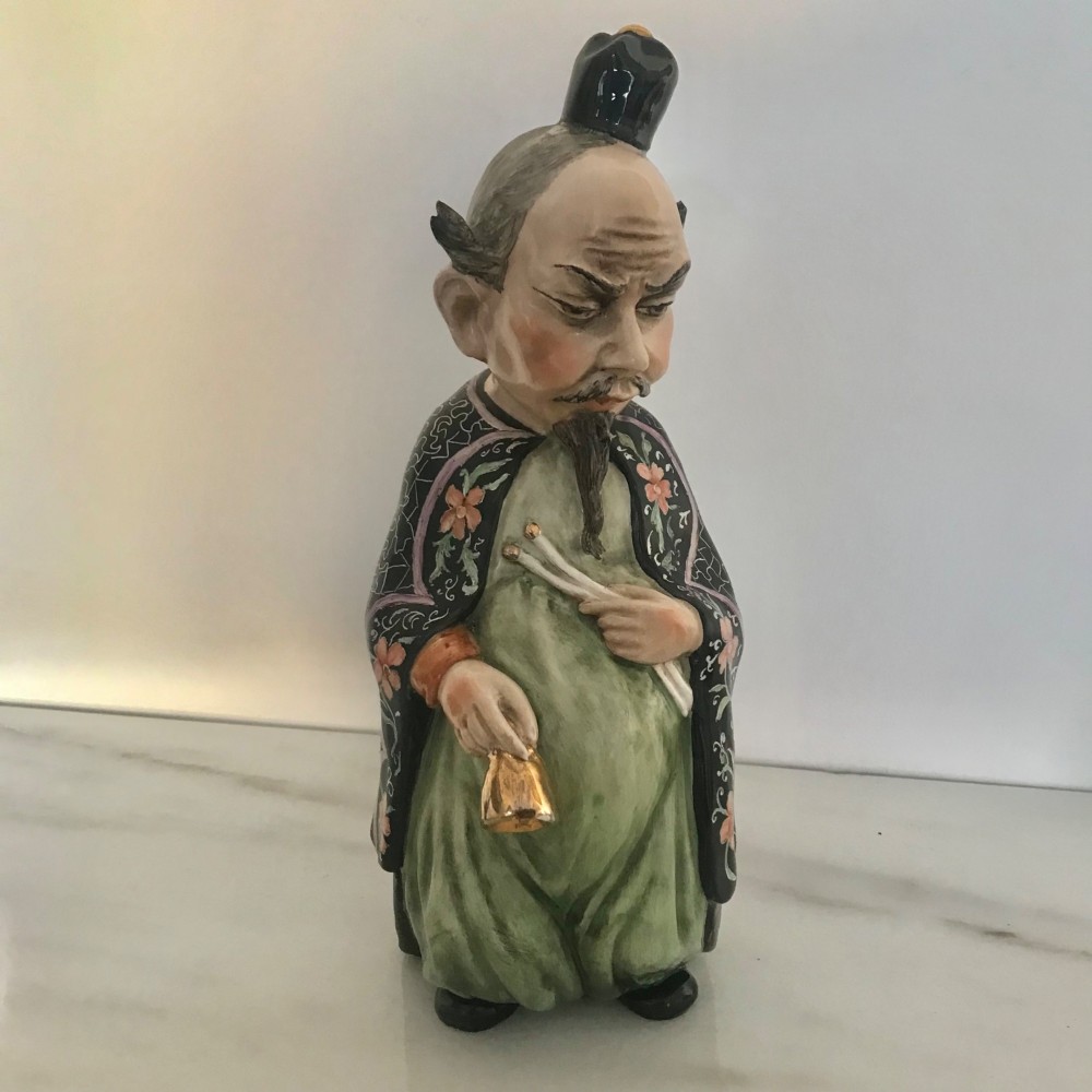 A Chinese ceramic figurine of a scholar wearing a black patterned robe and green garments, holding two scrolls and a small gold bell.