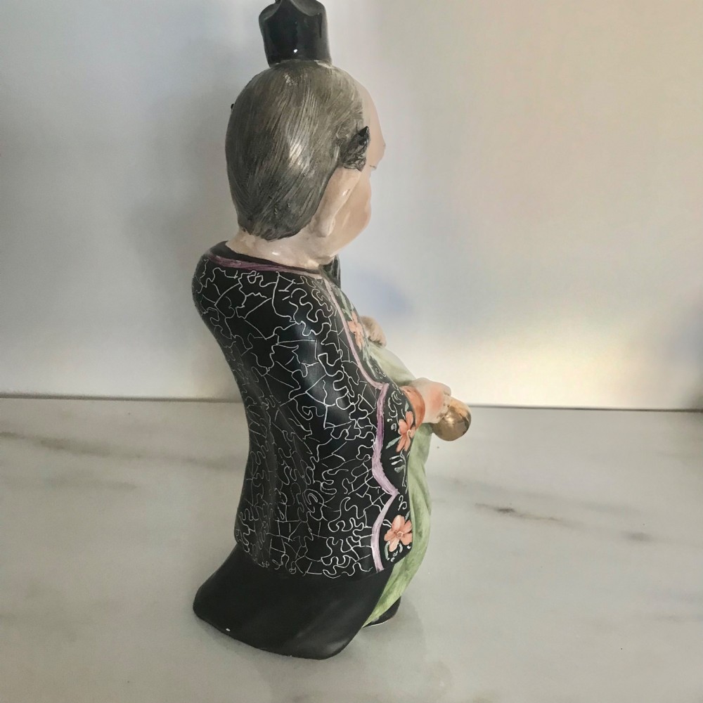 A Chinese ceramic figurine of a scholar wearing a black patterned robe and green garments, holding two scrolls and a small gold bell. View VI
