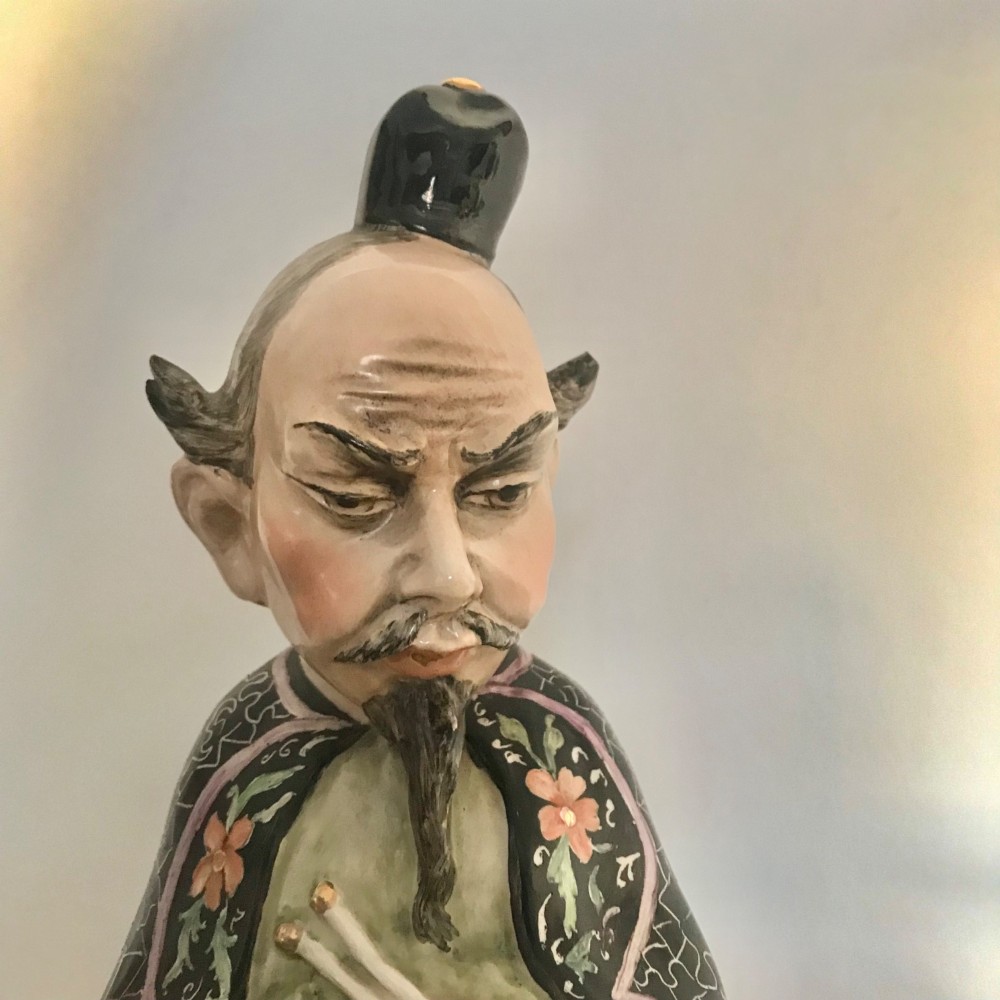 A Chinese ceramic figurine of a scholar wearing a black patterned robe and green garments, holding two scrolls and a small gold bell. View IX