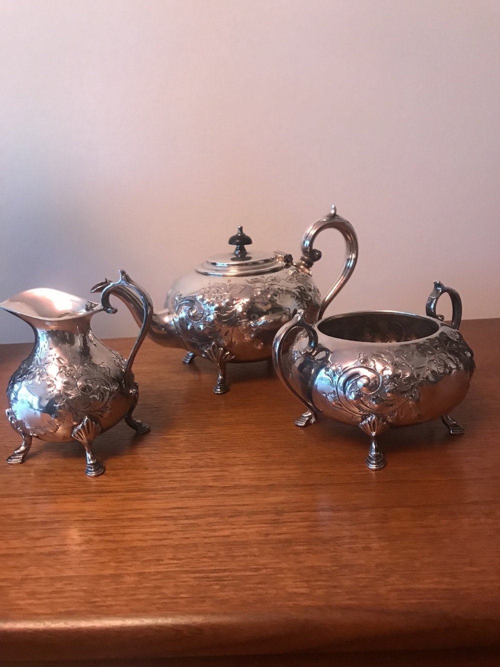 An Edwardian three-piece silver-plated tea set comprising a teapot, milk jug, and sugar bowl featuring floral chasing and scroll feet. View IV