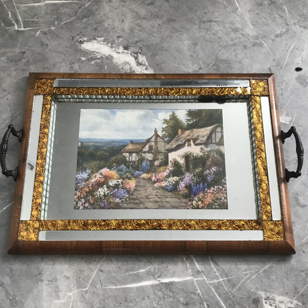 An Art Deco rectangular serving tray featuring a walnut veneer frame, metal side handles, and a mirrored glass surface with a central cottage landscape print framed by gold borders.