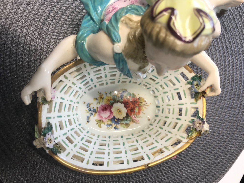 An 18th-century porcelain sweetmeat dish featuring a reclining female figure beside an openwork lattice basket adorned with hand-painted polychrome flowers and gilt trim. View X