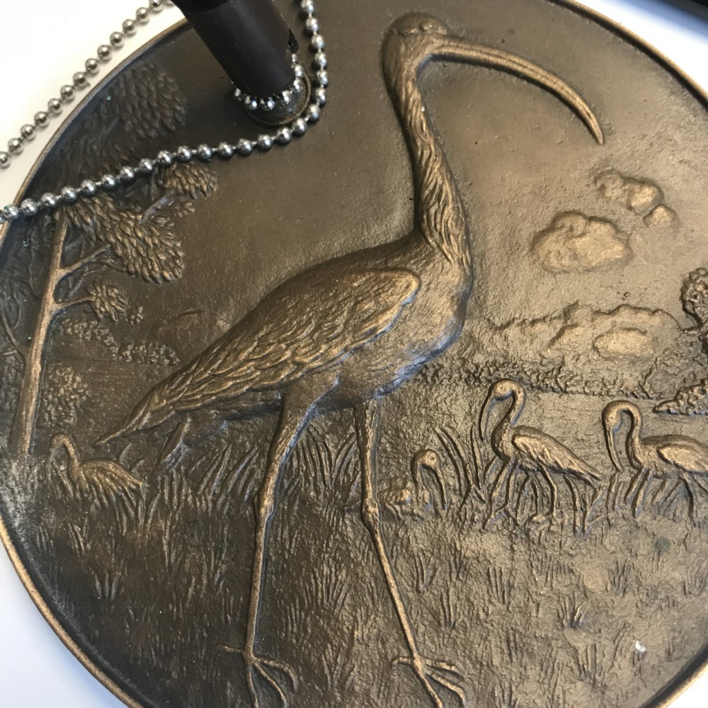 A round bronze medallion base featuring an embossed ibis bird in a landscape setting, complete with a black pen attached via a fine chain. View X