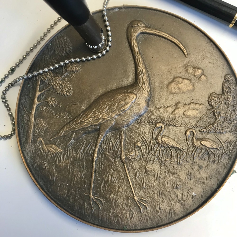 A round bronze medallion base featuring an embossed ibis bird in a landscape setting, complete with a black pen attached via a fine chain. View XI