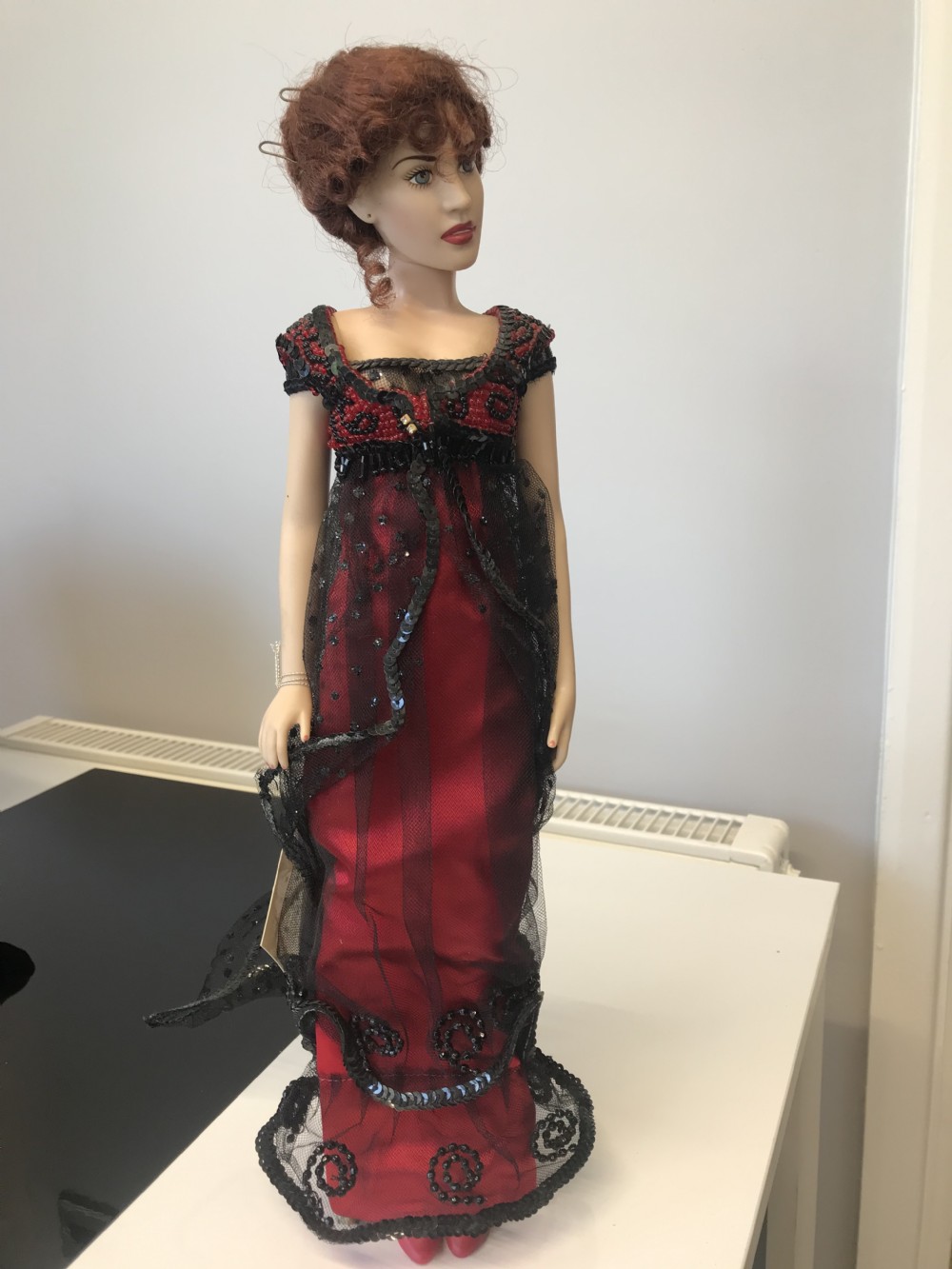A porcelain collector doll wearing a red evening gown with black beaded lace overlay and red hair styled in an updo.