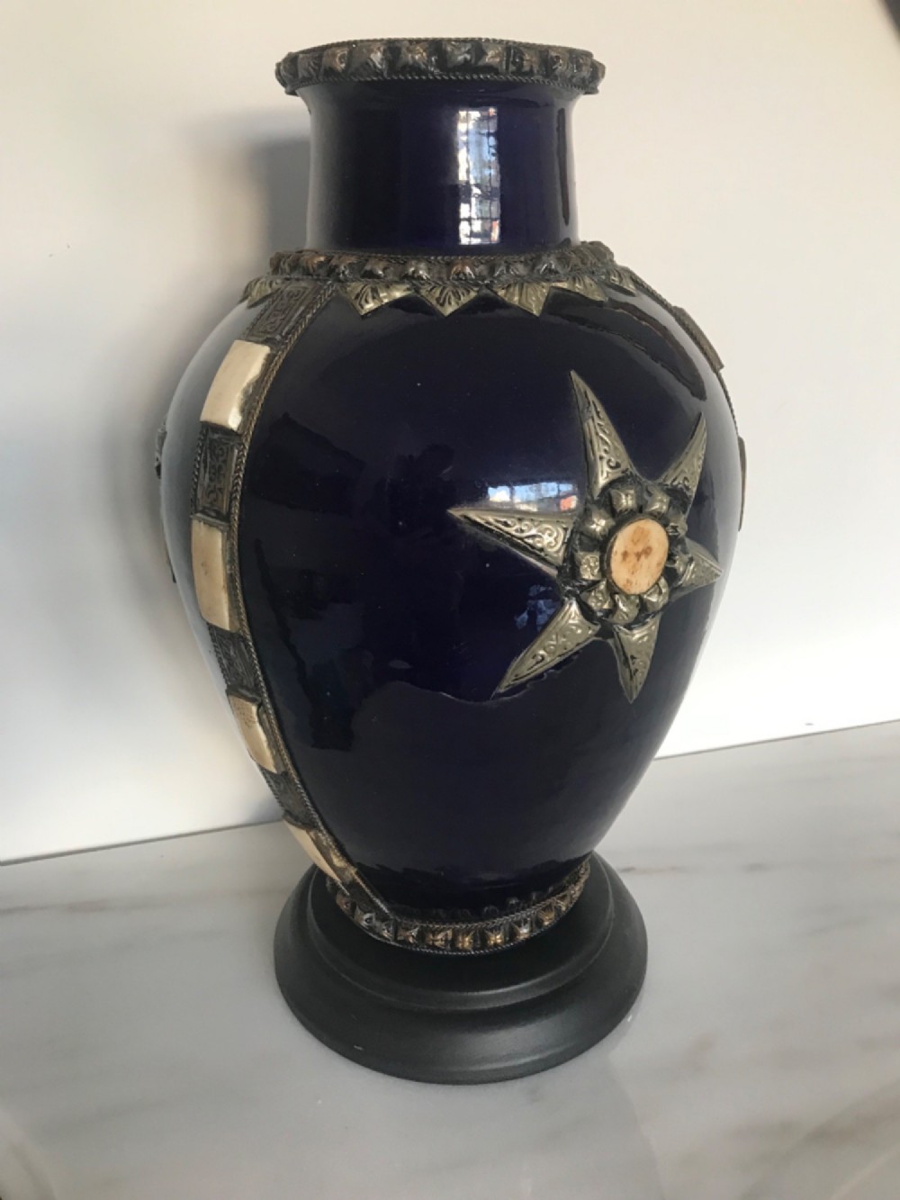 A cobalt blue glazed terracotta vase adorned with applied silver metalwork, a star motif, bone inlays, and mounted on a dark circular pedestal.