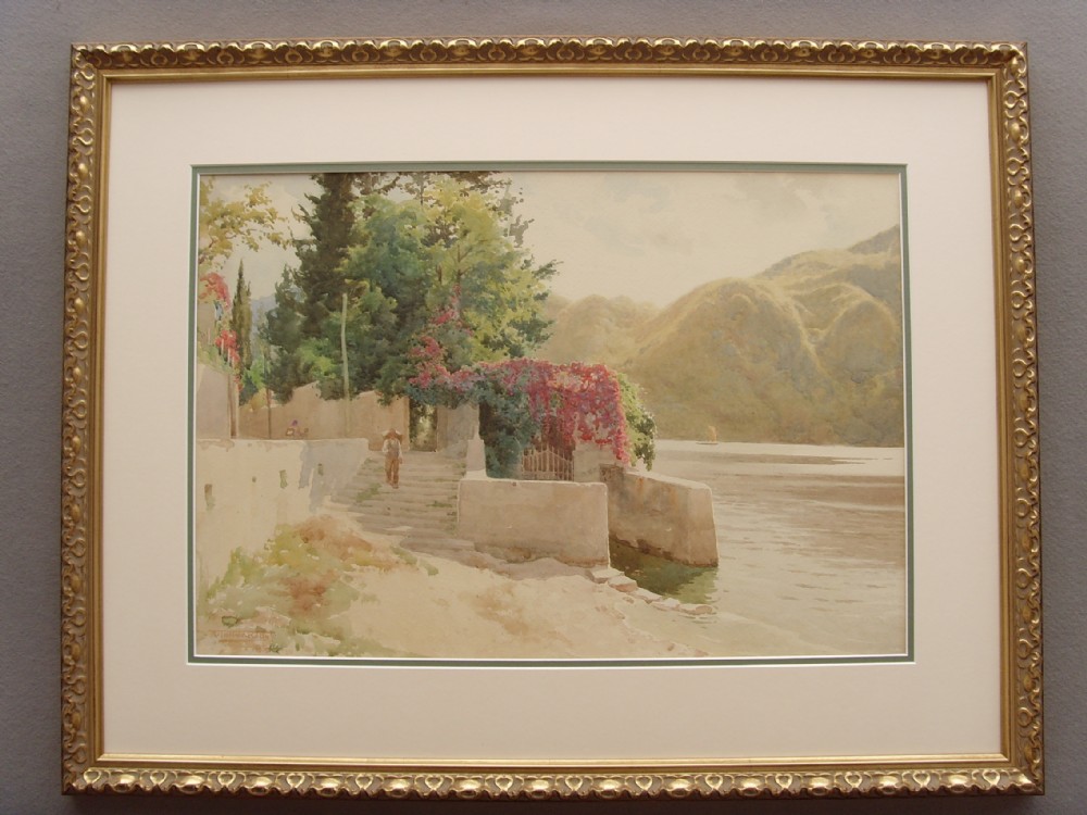 A framed Edwardian watercolour painting depicting a sunny Lake Como shore with lush green trees, stone steps, and distant mountains enclosed in an ornate gold gilt frame.