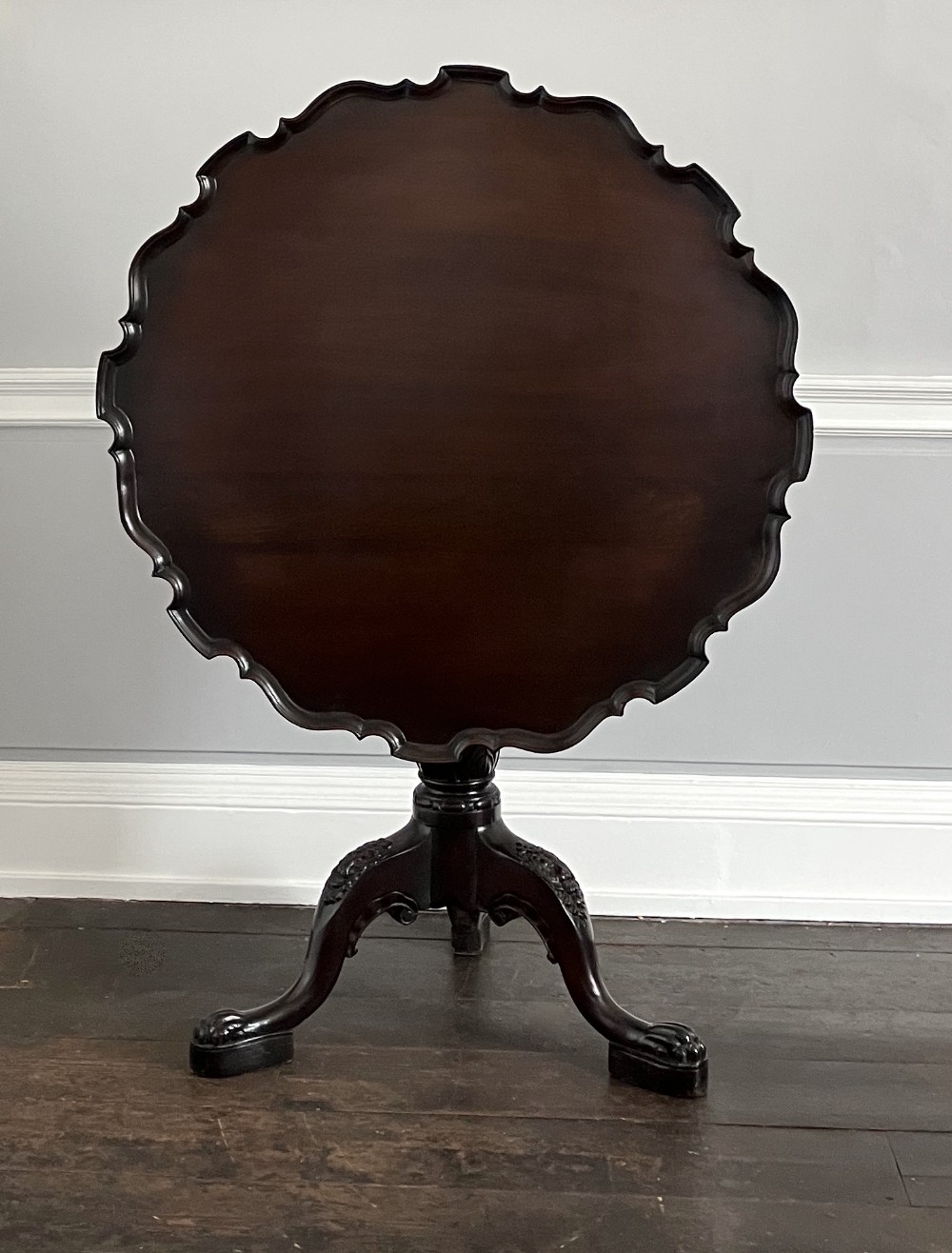A dark Georgian mahogany tilt-top tripod table with a carved piecrust edge displayed upright on a three-legged base.