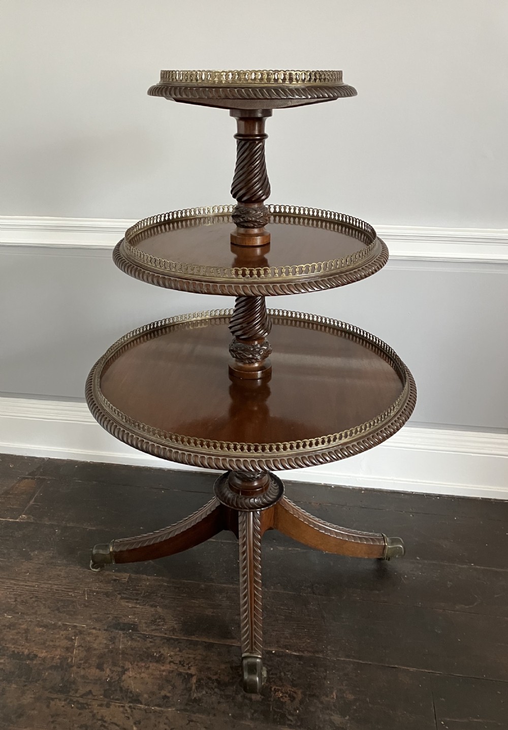 A three-tiered dark mahogany dumbwaiter table featuring pierced brass gallery rims, spiral-carved central supports, and splayed quadruped legs with brass castors.
