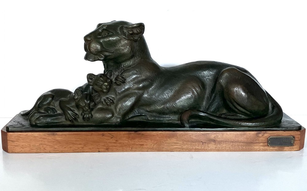 A dark patinated bronze sculpture depicting a reclining lioness alongside two cubs, mounted on a rectangular timber plinth. View II