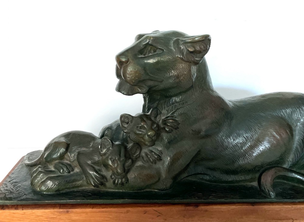 A dark patinated bronze sculpture depicting a reclining lioness alongside two cubs, mounted on a rectangular timber plinth. View III