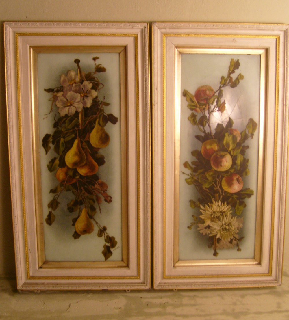 A pair of tall, narrow Victorian wall art panels depicting hand-painted fruit and floral arrangements set within painted cream and gilded wooden frames.