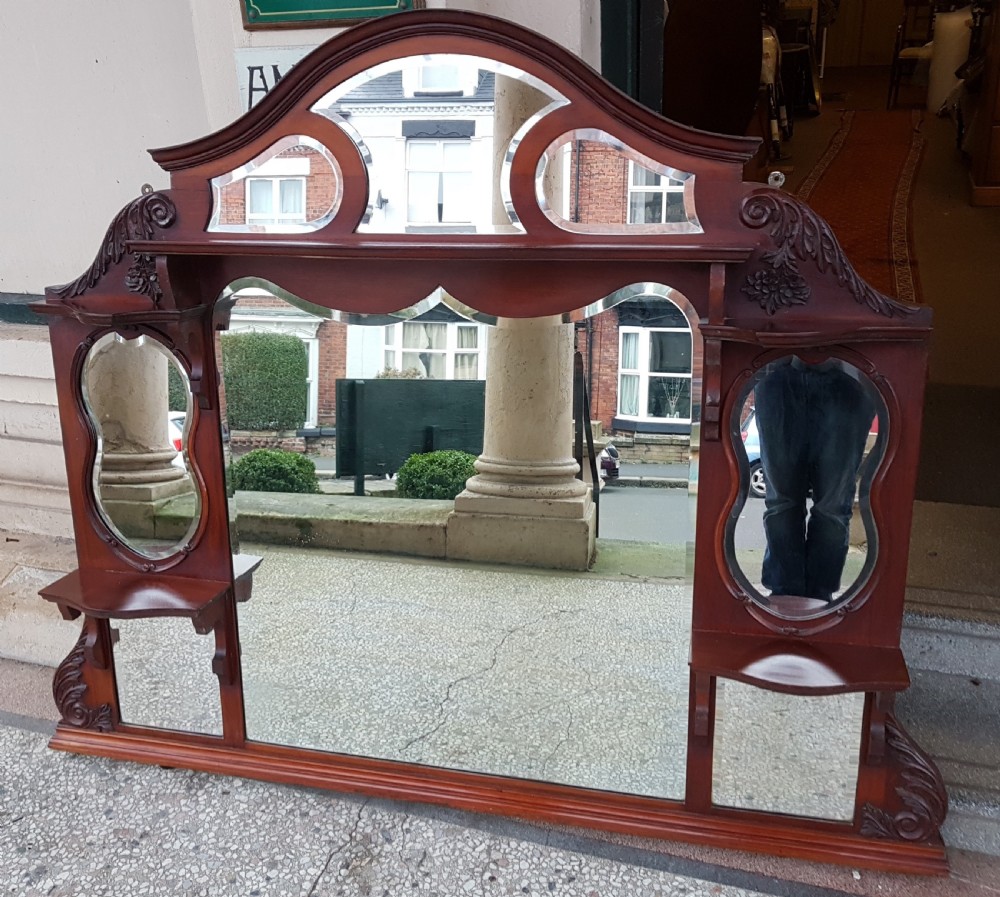 An Edwardian carved mahogany overmantel mirror featuring an arched crest, multiple bevelled glass panels, and integrated side display shelves.