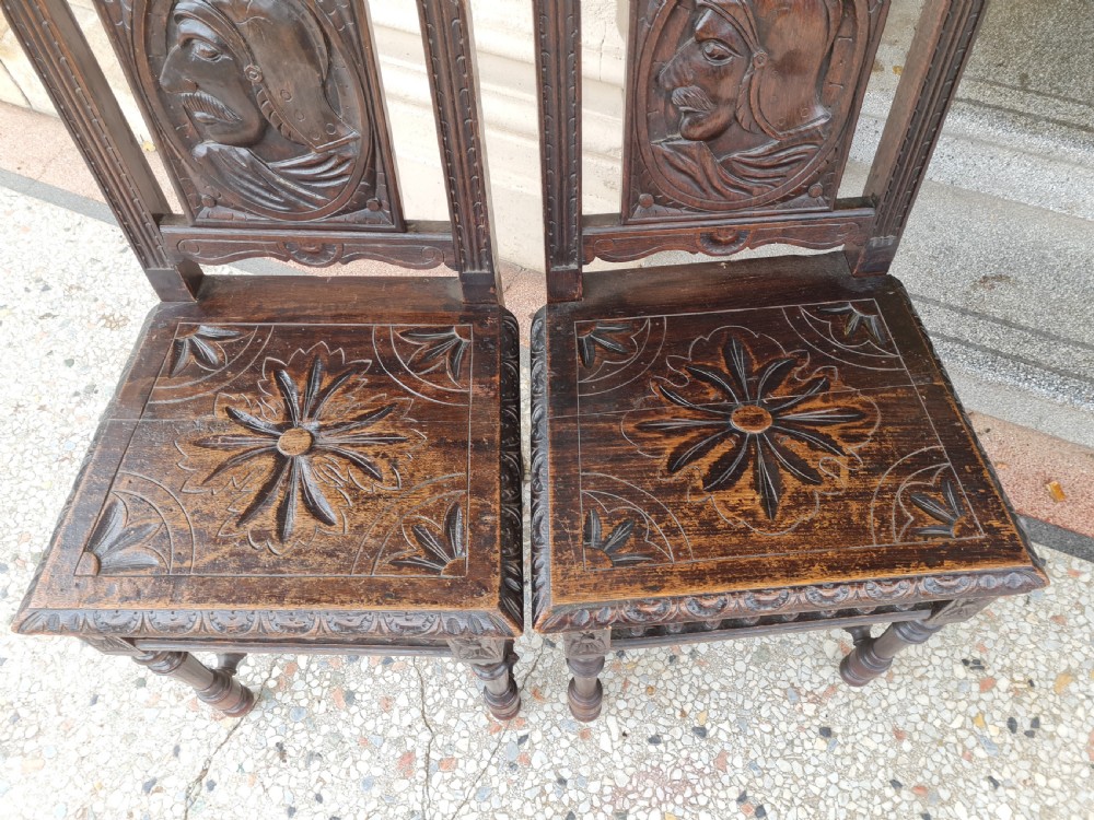 A pair of dark carved oak hall chairs featuring carved portrait medallions on the backrests and starburst carvings on the timber seats. View V