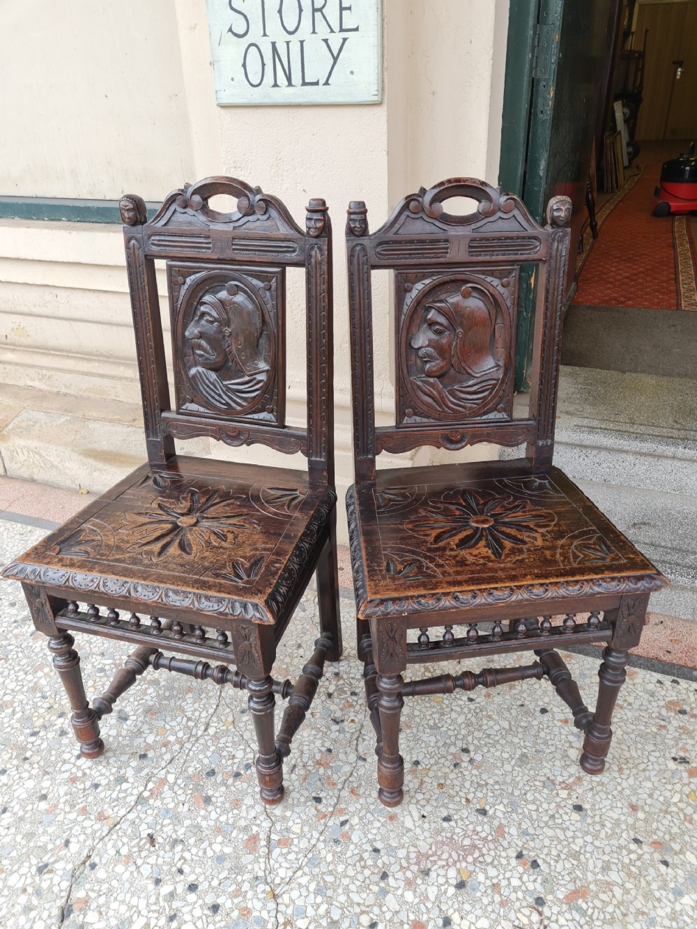 A pair of dark carved oak hall chairs featuring carved portrait medallions on the backrests and starburst carvings on the timber seats.