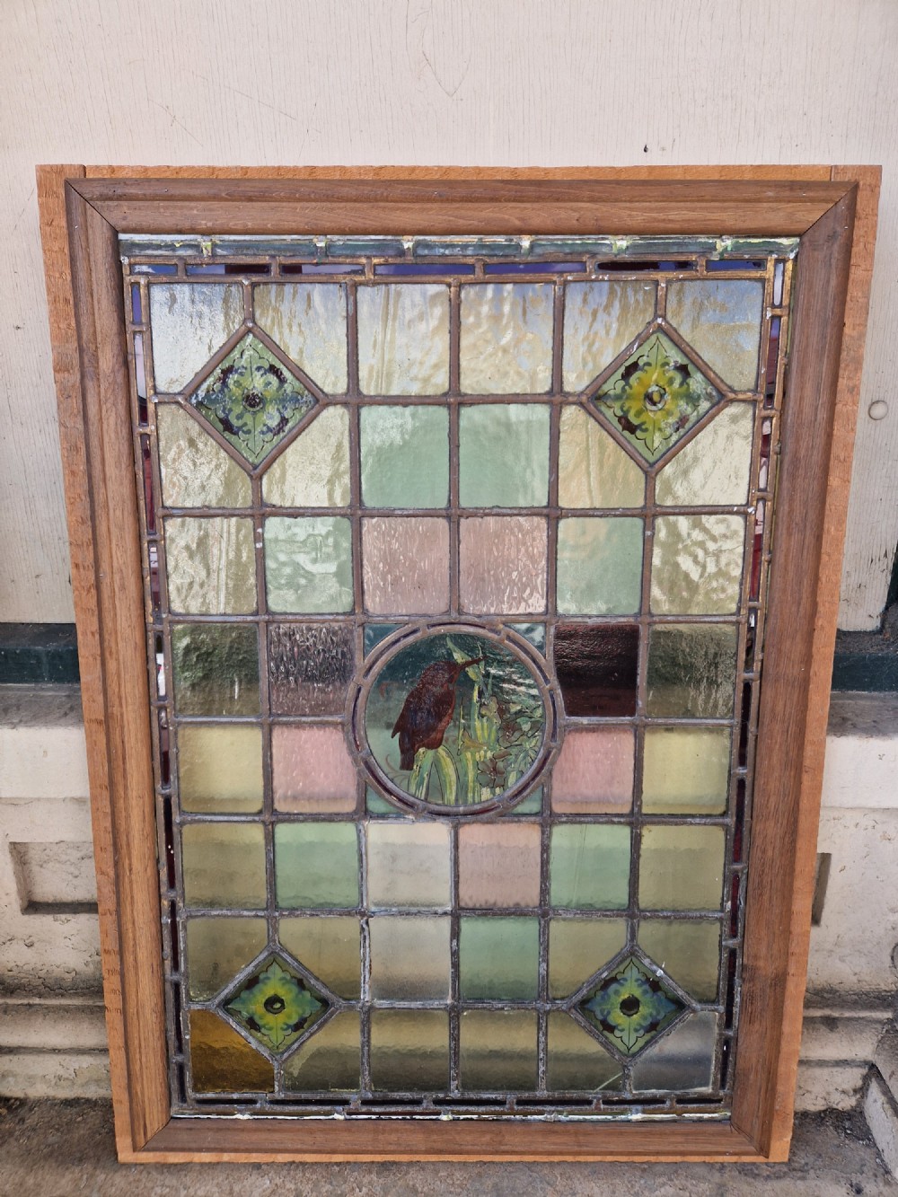 A rectangular Victorian leaded glass panel set in a timber frame, featuring pastel geometric panes, four decorative corner inserts, and a central roundel depicting a hand-painted bird.