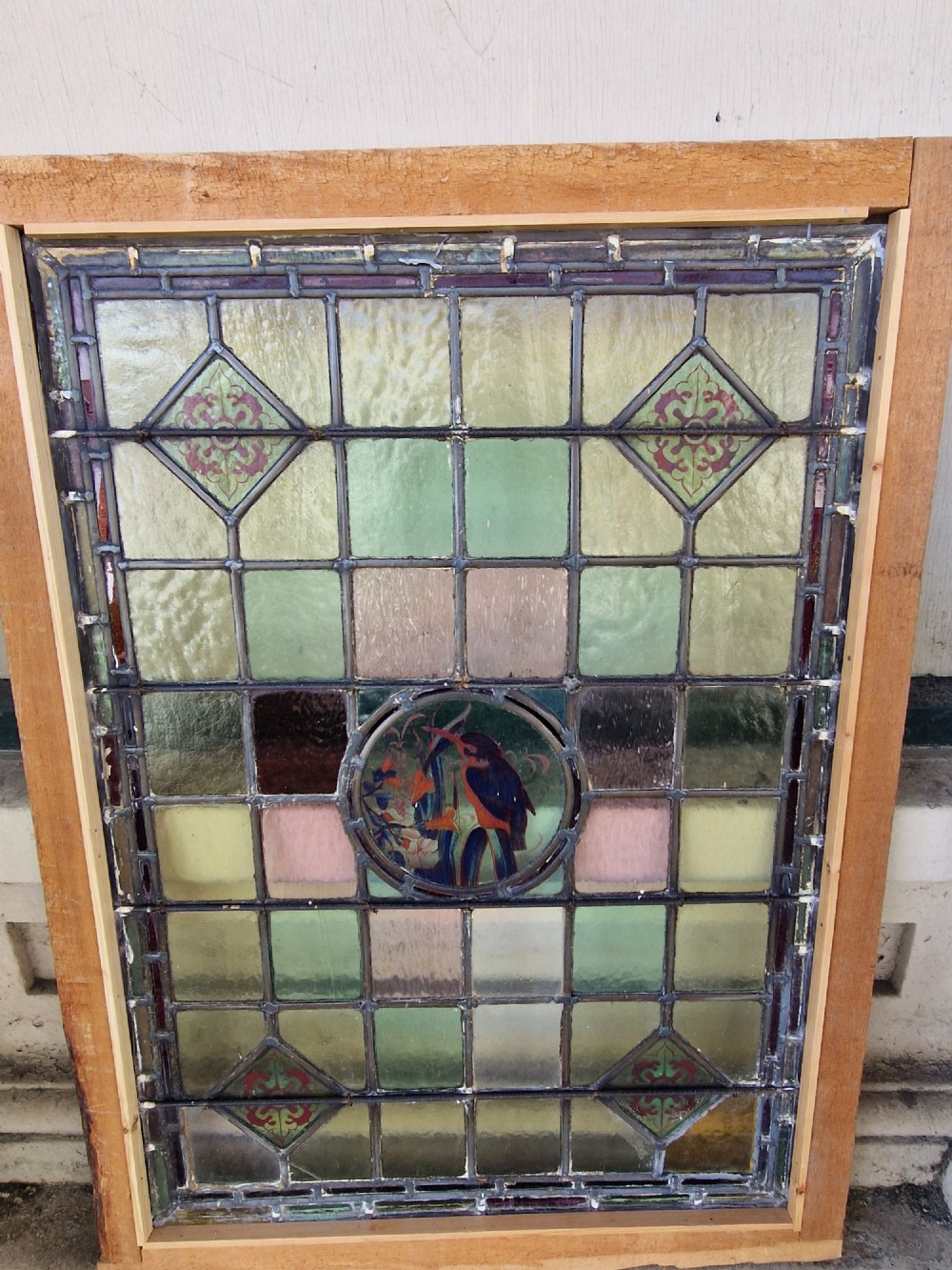 A rectangular Victorian leaded glass panel set in a timber frame, featuring pastel geometric panes, four decorative corner inserts, and a central roundel depicting a hand-painted bird. View VII