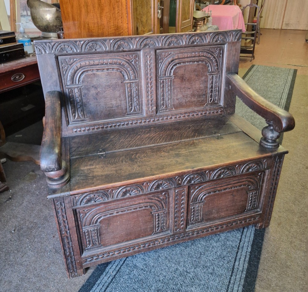 A dark carved oak settle bench with a panelled back, sculpted armrests, and a hinged lid seat above a carved front storage box.