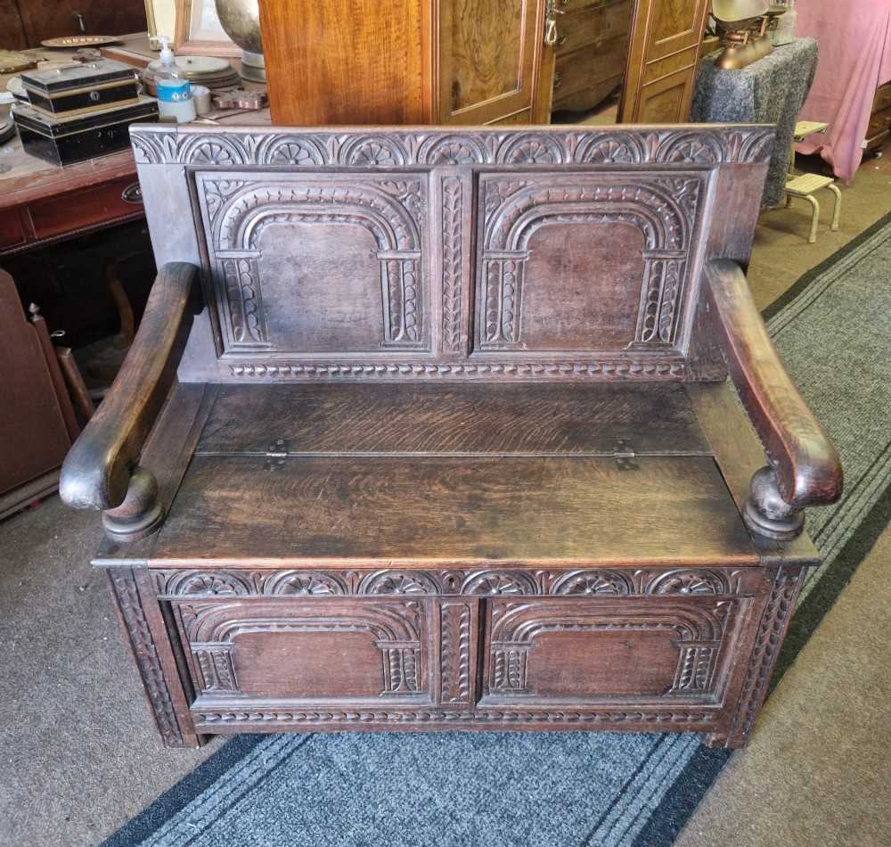 A dark carved oak settle bench with a panelled back, sculpted armrests, and a hinged lid seat above a carved front storage box. View II