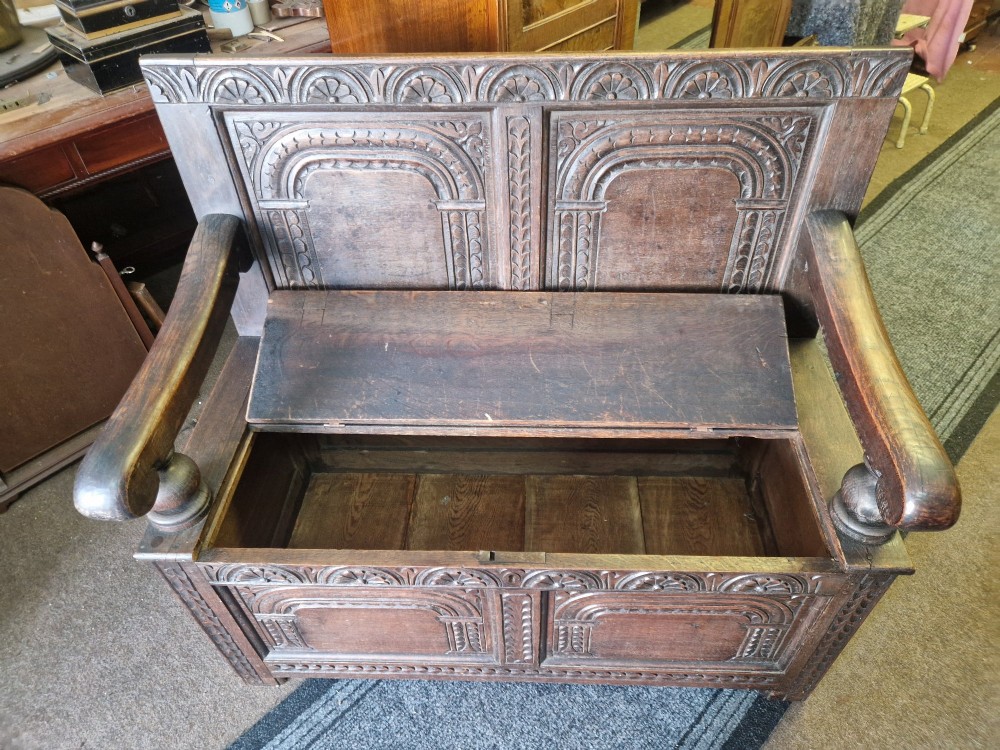 A dark carved oak settle bench with a panelled back, sculpted armrests, and a hinged lid seat above a carved front storage box. View III