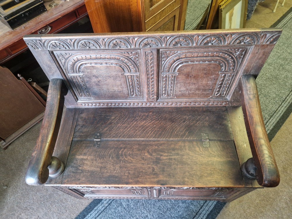 A dark carved oak settle bench with a panelled back, sculpted armrests, and a hinged lid seat above a carved front storage box. View IV