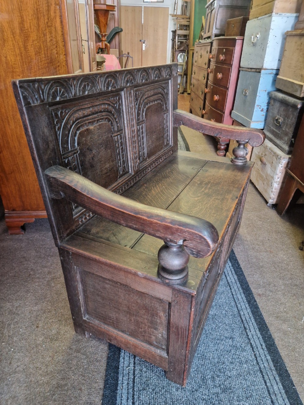 A dark carved oak settle bench with a panelled back, sculpted armrests, and a hinged lid seat above a carved front storage box. View V
