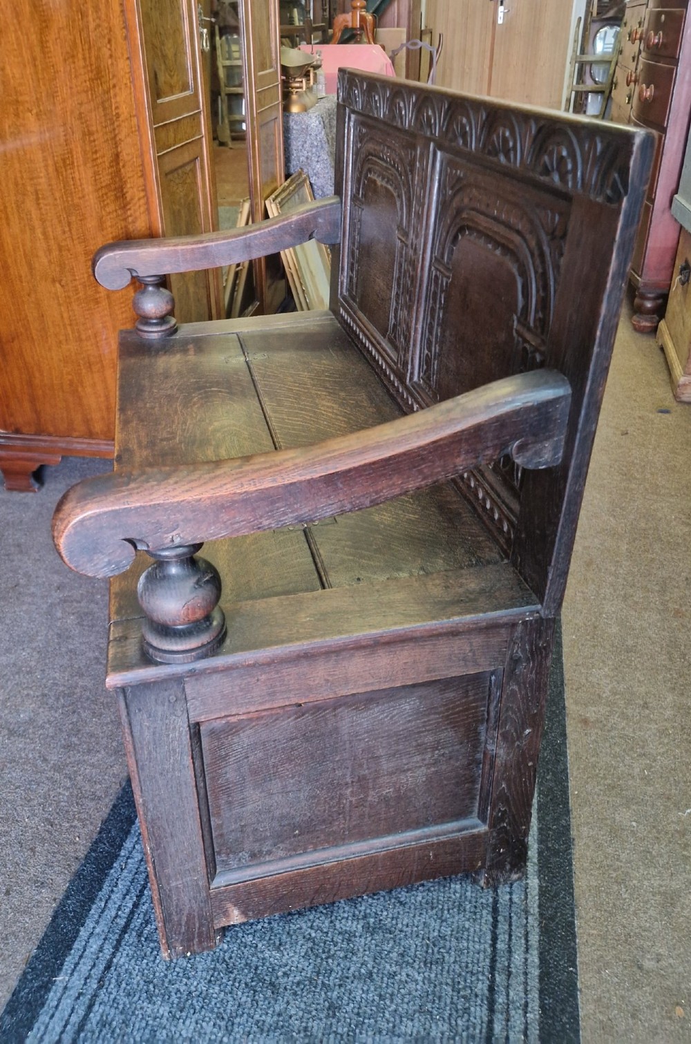 A dark carved oak settle bench with a panelled back, sculpted armrests, and a hinged lid seat above a carved front storage box. View VI