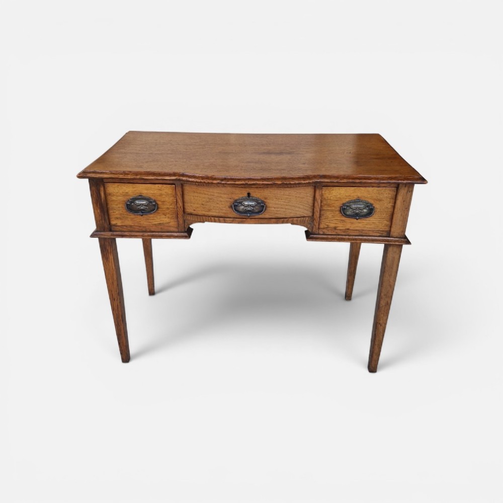 An Edwardian solid oak writing desk featuring three frieze drawers with metal drop handles, set on four square tapered legs against a neutral background.