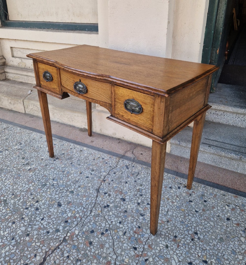 An Edwardian solid oak writing desk featuring three frieze drawers with metal drop handles, set on four square tapered legs against a neutral background. View II