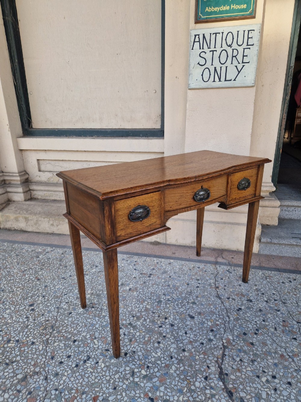 An Edwardian solid oak writing desk featuring three frieze drawers with metal drop handles, set on four square tapered legs against a neutral background. View IV