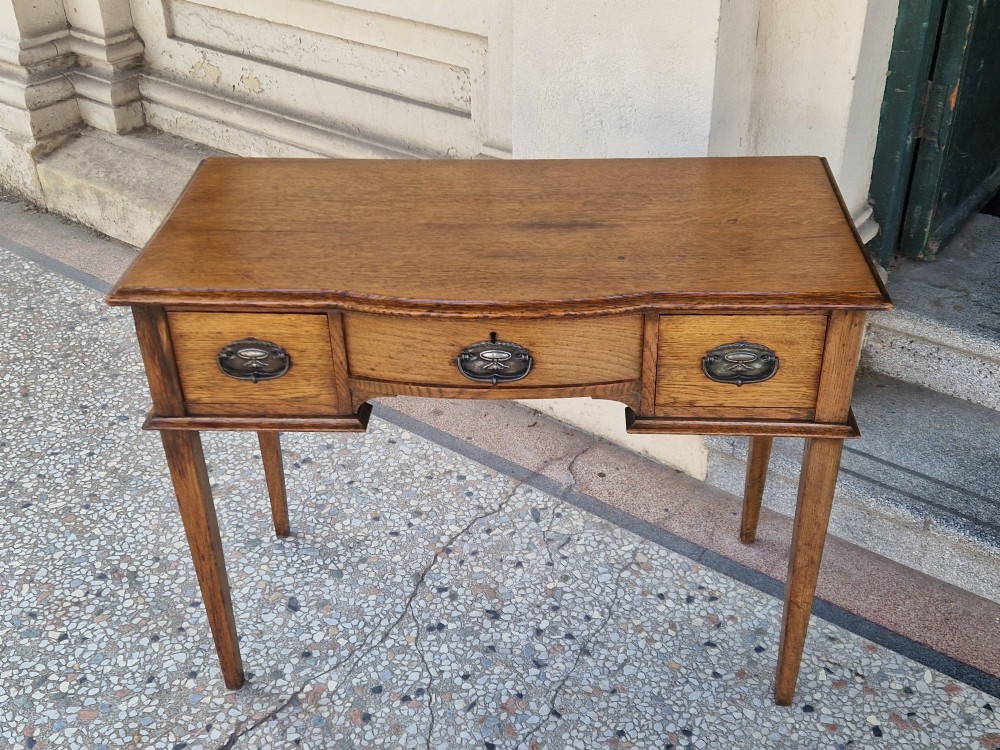 An Edwardian solid oak writing desk featuring three frieze drawers with metal drop handles, set on four square tapered legs against a neutral background. View V