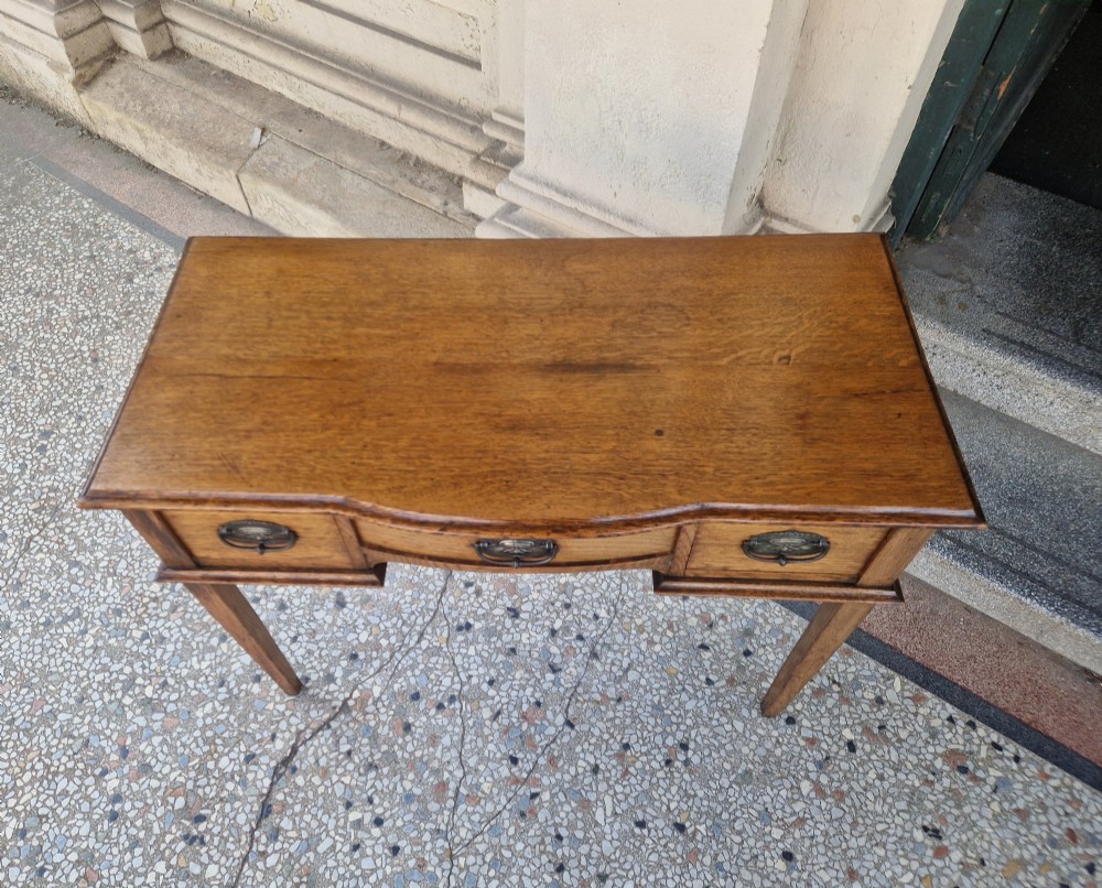 An Edwardian solid oak writing desk featuring three frieze drawers with metal drop handles, set on four square tapered legs against a neutral background. View VI