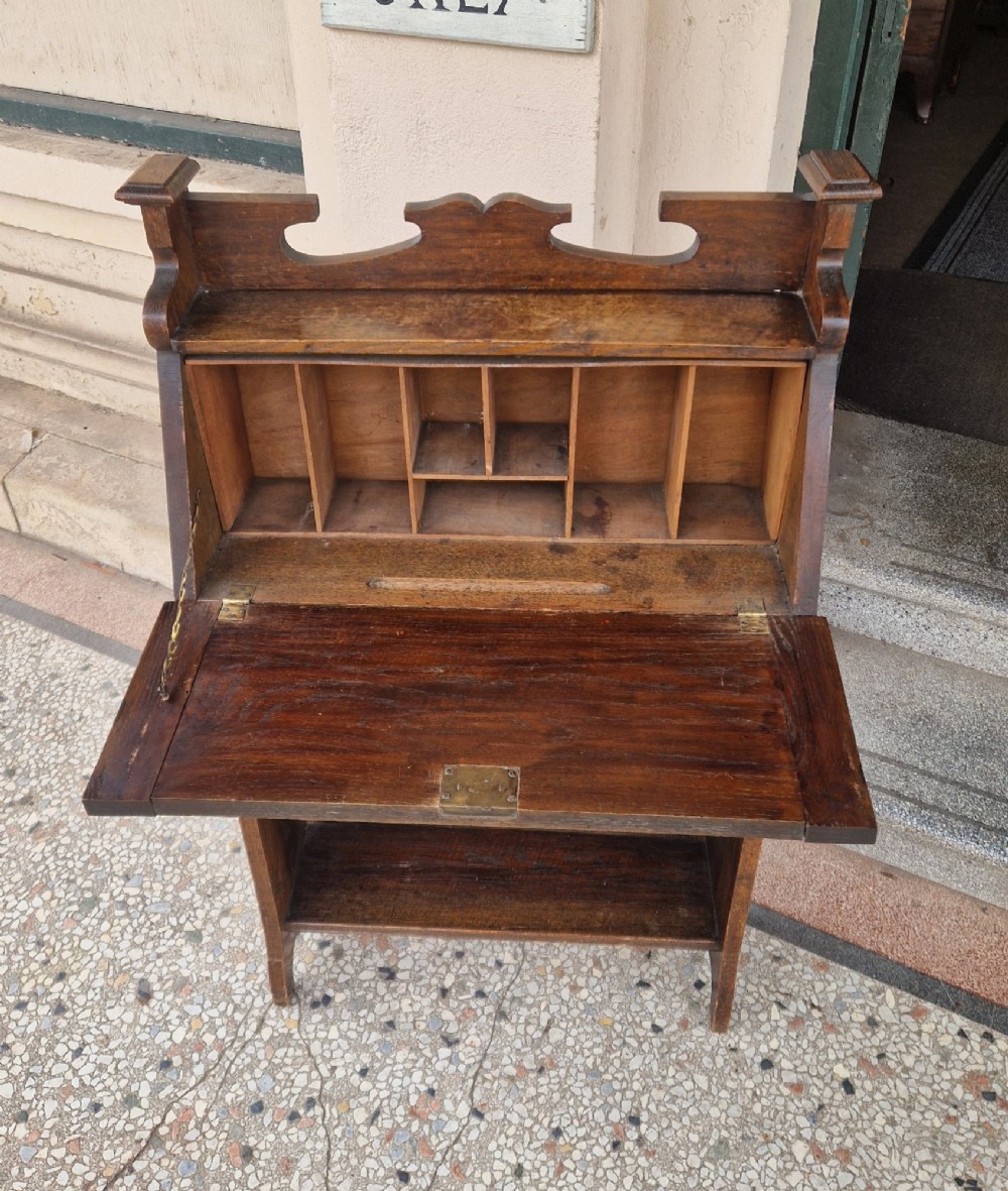 An Arts and Crafts oak writing bureau with a slant-front lid, copper strap hinges, a carved top cresting rail, and two open shelves below. View II