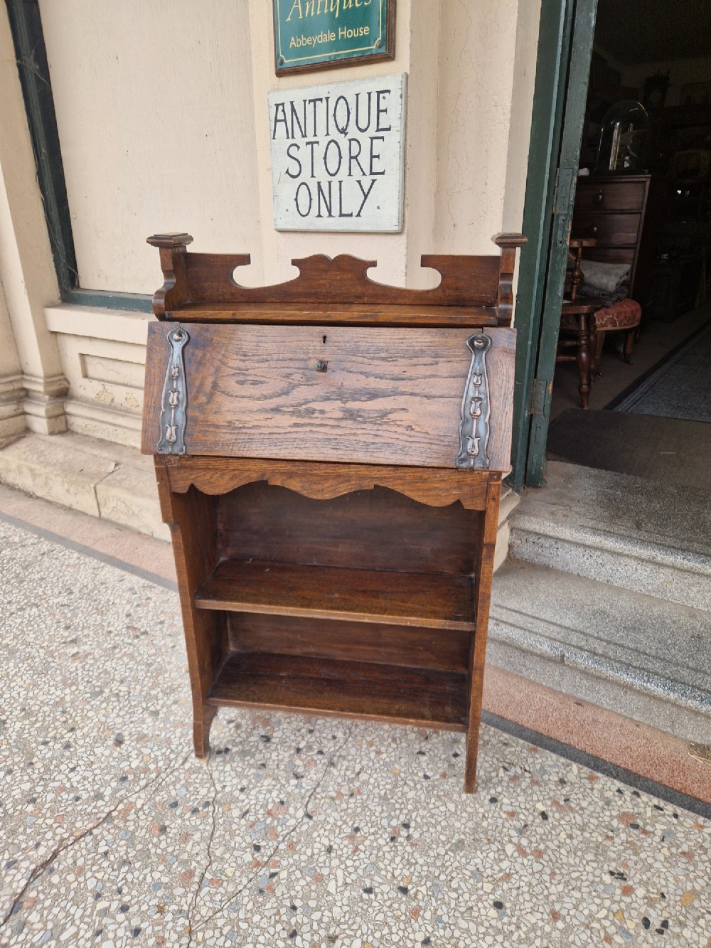 An Arts and Crafts oak writing bureau with a slant-front lid, copper strap hinges, a carved top cresting rail, and two open shelves below. View V