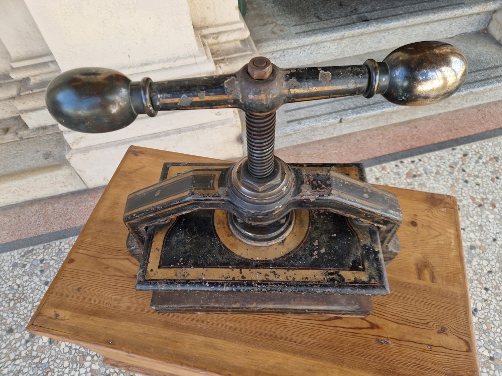 A heavy black cast iron book press featuring a central screw mechanism with oversized rounded handles and painted gold line detailing. View III