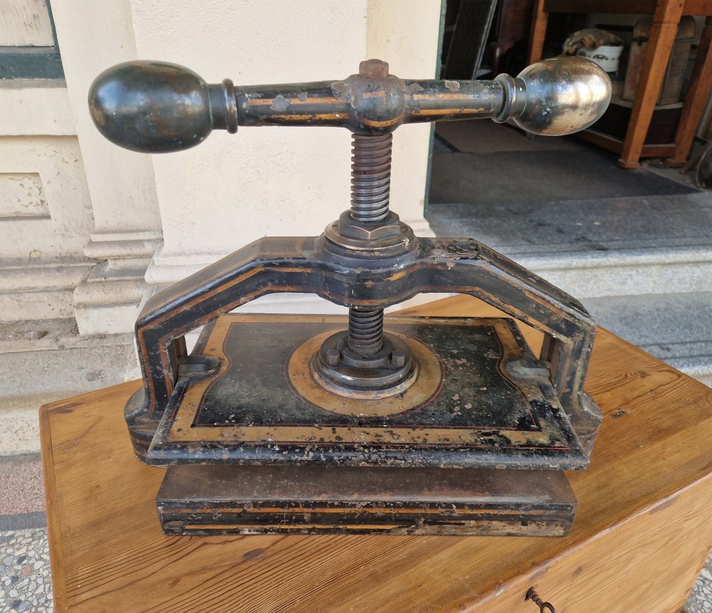 A heavy black cast iron book press featuring a central screw mechanism with oversized rounded handles and painted gold line detailing. View V
