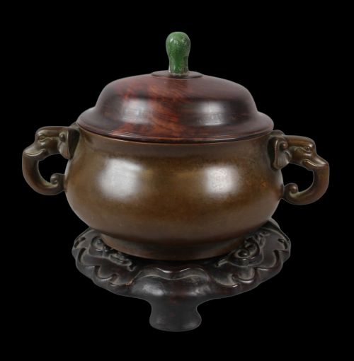 fig 1. A rounded dark bronze Chinese censer with elephant head handles, topped with a polished wood lid featuring a green jade finial, resting on a carved hardwood stand.