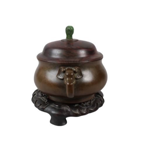 fig 2. A rounded dark bronze Chinese censer with elephant head handles, topped with a polished wood lid featuring a green jade finial, resting on a carved hardwood stand.