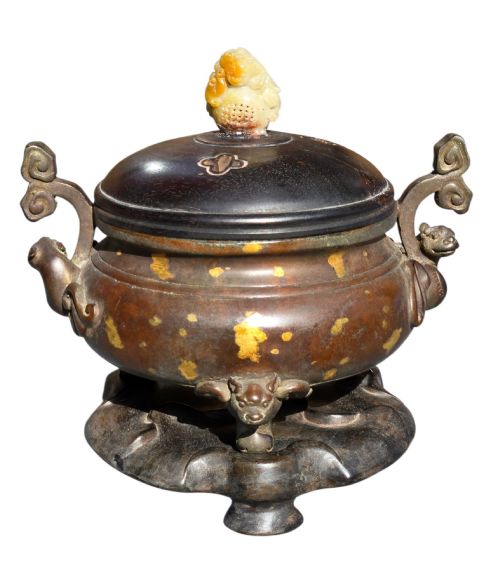 fig 2. An 18th-century Chinese gilt-splashed bronze tripod censer featuring scroll handles, animal-mask legs, a dark hardwood cover topped with a carved jade finial, and a fitted bronze stand.