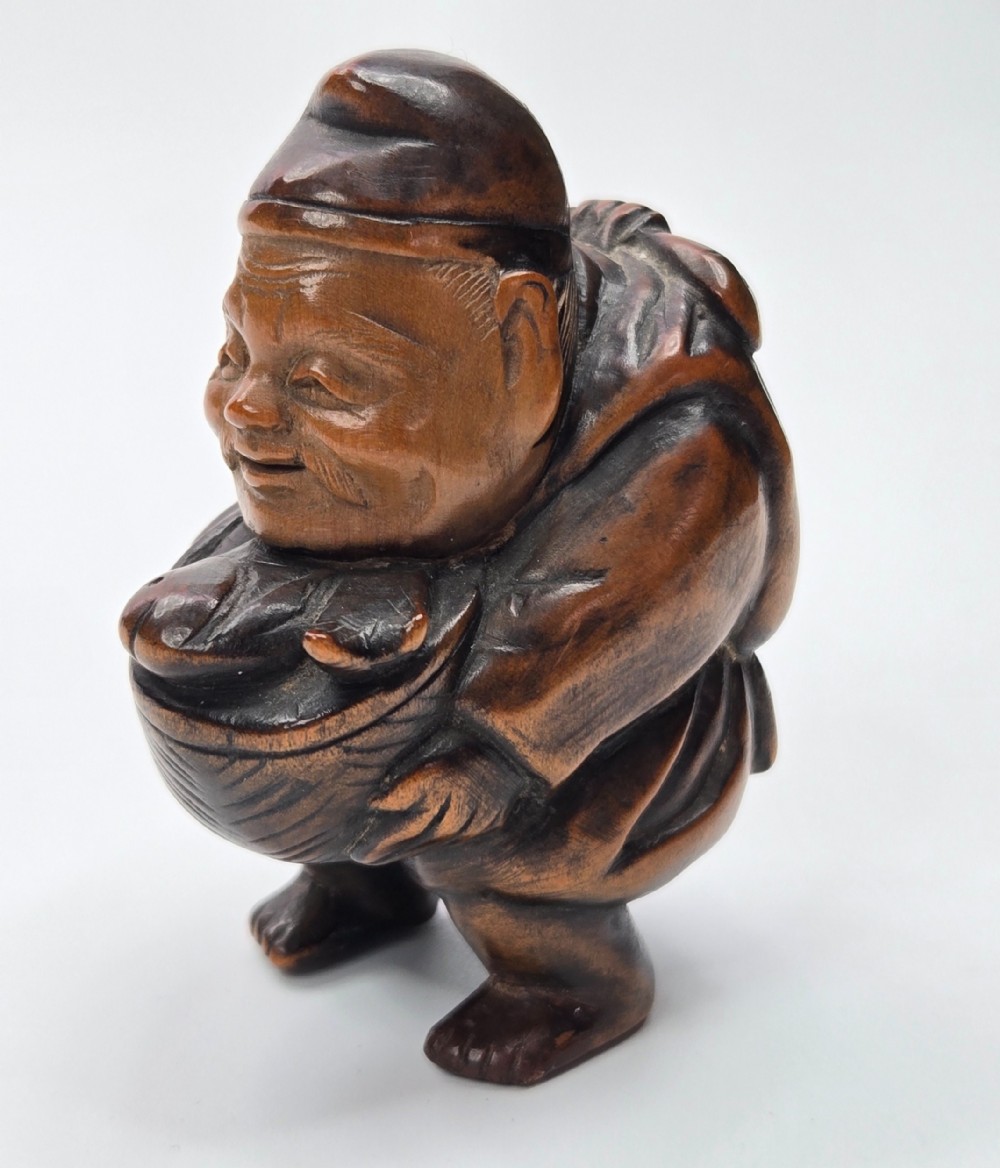 A carved dark wood Japanese figurine depicting Ebisu holding a basket containing a sea bream fish.