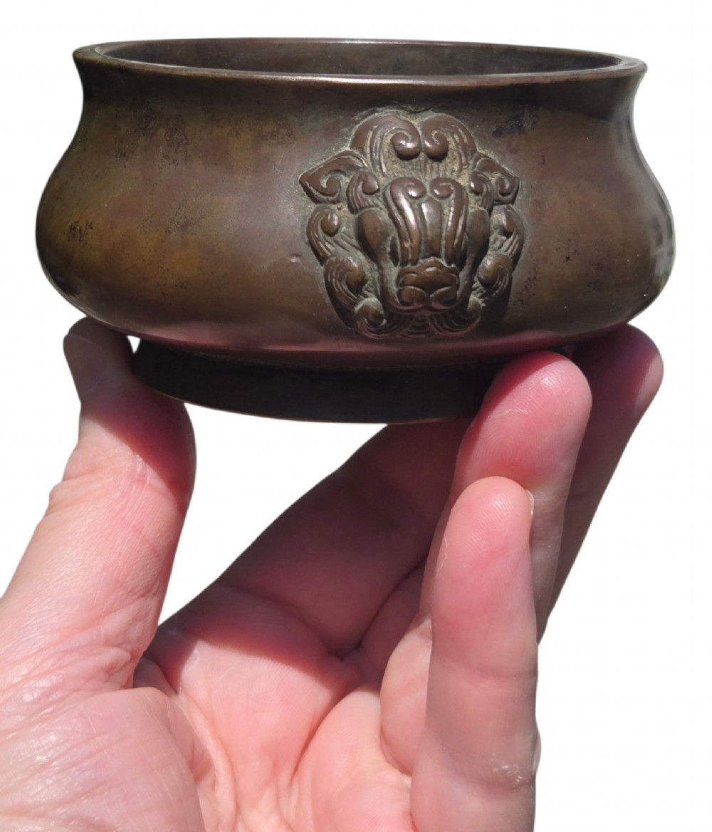 A cast bronze censer featuring a flared rim, compressed globular body, low circular foot, and cast relief taotie mask handles on the side. View IX