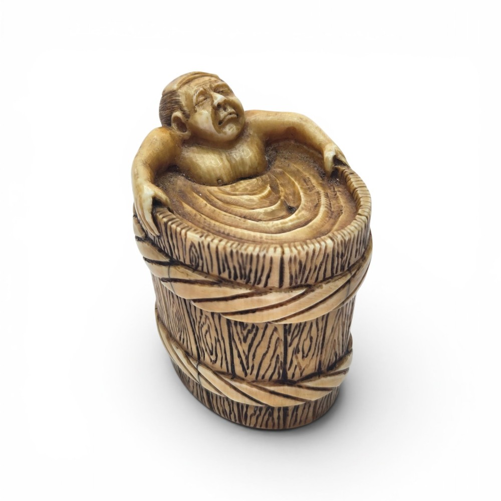 A small carved stag antler netsuke depicting a man sitting inside a wooden bath tub with carved water ripples.
