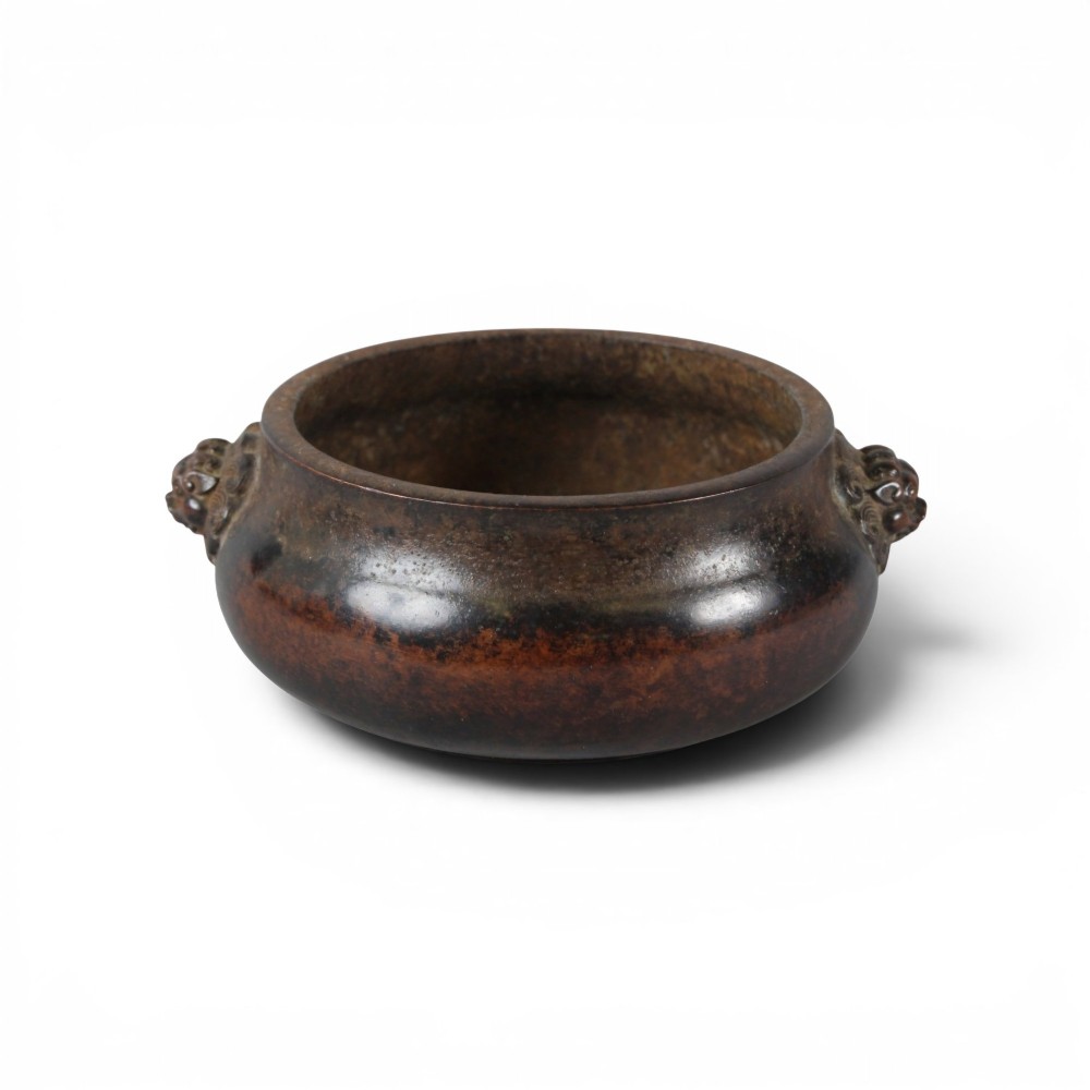 A heavy dark brown cast bronze incense burner featuring a low compressed circular form decorated with relief beast mask handles. View III