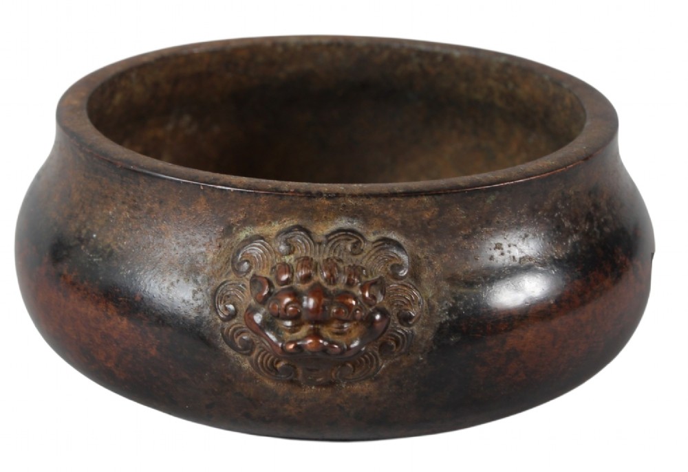 A heavy dark brown cast bronze incense burner featuring a low compressed circular form decorated with relief beast mask handles. View V