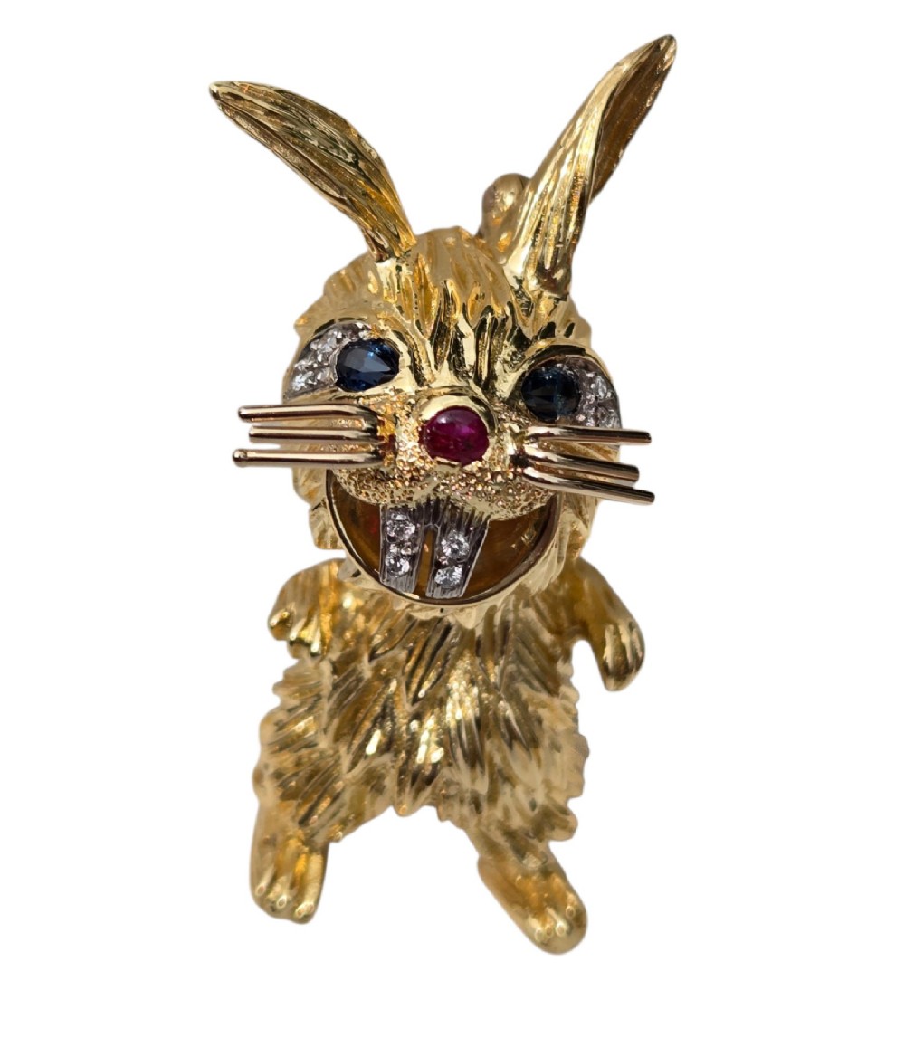 An 18ct yellow gold rabbit brooch showcasing textured fur detailing, a ruby nose, diamond-set teeth, and sapphire eyes. View VI