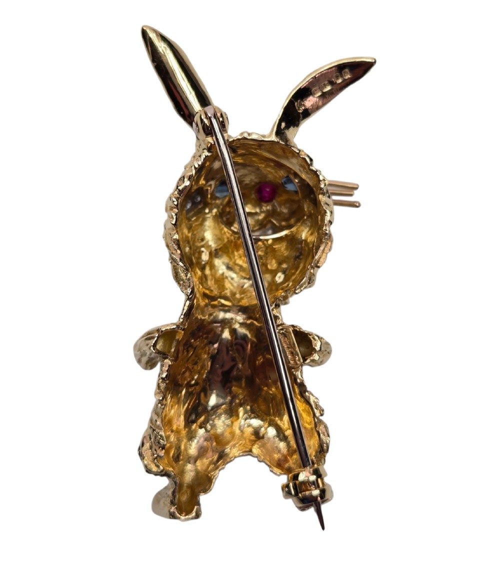 An 18ct yellow gold rabbit brooch showcasing textured fur detailing, a ruby nose, diamond-set teeth, and sapphire eyes. View X